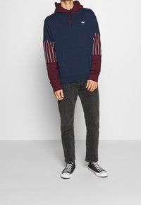 Marinblå hoodie med vinröda ärmar som har vita ränder, framficka och liten logotyp. Bärs med svarta jeans och sneakers.