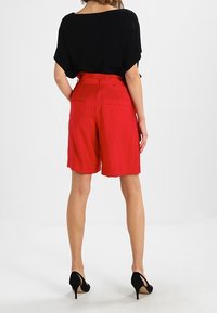 Rote Shorts mit taillierter Passform, bis zur Mitte des Oberschenkels reichend, ausgestattet mit zwei Gesäßtaschen, kombiniert mit einem schwarzen Oberteil und schwarzen High Heels.