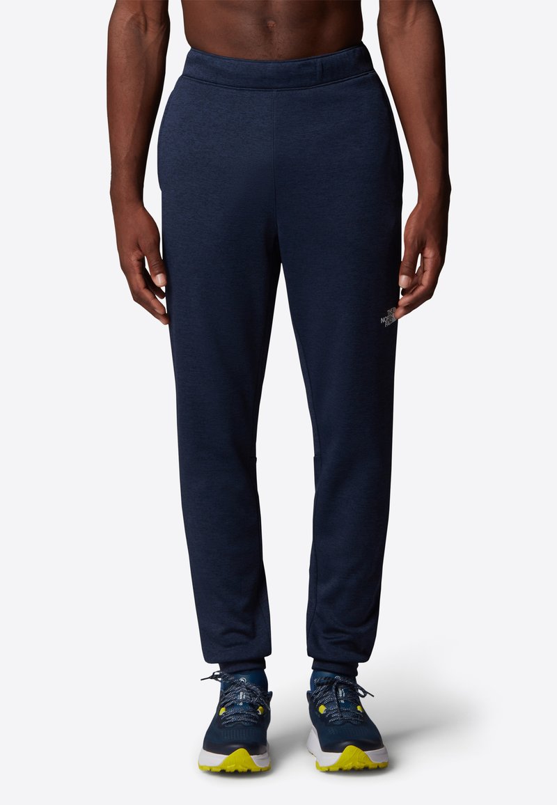 Marinblå träningsbyxor med avslappnad passform, elastisk midja och sidofickor. Kombineras med mörka skor med gula sulor och reflekterande snören.