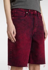 Pantalones cortos de mezclilla roja con una textura desgastada, de cintura alta y dobladillo deshilachado. Presentan bolsillos frontales y cierres de botón de metal.