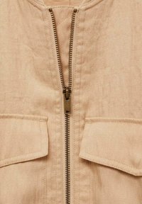Beżowa kurtka z dwoma naszywanymi przednimi kieszeniami, posiadająca centralny mosiężny zamek błyskawiczny, teksturowaną tkaninę i okrągły dekolt.