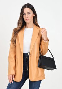 Blazer in finta pelle color senape con rever a notched, tasche frontali, indossato sopra una maglietta bianca e jeans a vita alta blu; borsa nera dal design strutturato.
