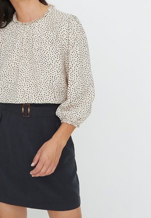 Kvinde iført en cremefarvet bluse med sorte polkaprikker og et sort skørt med en rektangulær bæltespænde, stående med den ene hånd synlig.