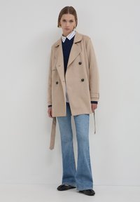 Frau in beigem doppelt geknöpftem Mantel, marineblauem Pullover über gestreiftem Hemd, hellblauen Schlagjeans und schwarzen Schuhen, die vor einer weißen Wand steht.