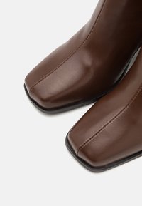 Bottes chevilles en cuir marron avec une texture lisse, un design à bout carré et des détails de couture contrastants. Semelle en caoutchouc noire et style minimaliste.