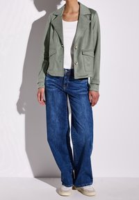 Light green suede blazer with a lapel collar, double-breasted buttons, and front pockets, paired with blue wide-leg jeans and light shoes.