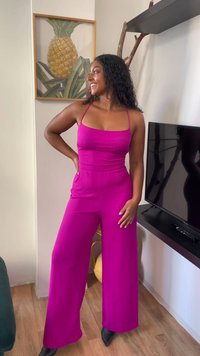 Mulher em um macacão sem mangas magenta, de pé dentro de casa perto de uma televisão e uma obra de arte de abacaxi, sorrindo e olhando para o lado.