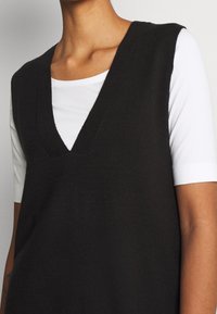 Haut sans manches en maille noire avec un col en V, superposé sur une chemise blanche à manches courtes. La texture est douce avec une finition lisse.