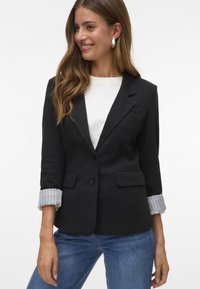 Črn blazer s krojem po meri, ki ima dva sprednja žepa in zavihane rokave z belimi črtami. Gladka tekstura tkanine.