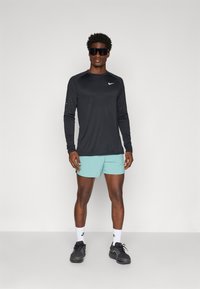 Sort langærmet træningstrøje med tekstureret design, lyseblå træningsshorts, sorte sneakers og mørke solbriller.