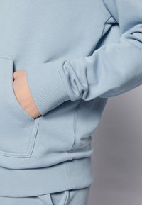 Sudadera de color azul claro con puños y dobladillo acanalados; presenta un bolsillo frontal y una tela suave y texturizada. Vista en primer plano de la manga y el bolsillo.