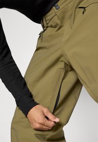 Pantalons de randonnée vert olive avec une texture lisse, poches zippées et un design ajusté. Accents noirs sur les zips et la ceinture.