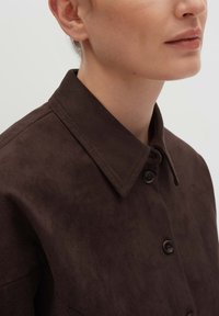 Braune Wildlederbluse mit einer Knopfleiste, klassischem Kragen und glatter Textur. Auffällige Details sind die dunklen Knöpfe und der dezente Glanz.