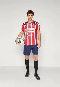 Rot-weißer gestreifter Fußballtrikot mit blauen Shorts, schwarzen Socken und schwarzen Fußballschuhen. Der Spieler hält einen schwarz-weißen Fußball.