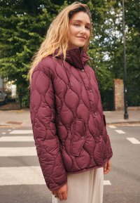 Veste matelassée bordeaux avec un col montant, deux poches avant et des fermetures par pressions. Texture douce et design rembourré pour la chaleur.