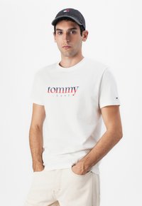 Hvit bomulls t-skjorte med "tommy JEANS" logo i rødt og marineblått, rund hals, korte ermer; kombinert med lysebeige bukser.