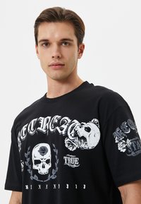 Schwarzes Baumwoll-T-Shirt mit einem großen Grafikdesign, das Schädel, Rosen und weißen Text zeigt, vor einem dunklen Hintergrund.