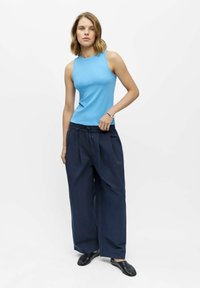 Mujer joven con camiseta sin mangas azul claro, pantalones sueltos azul oscuro y zapatos negros, de pie contra un fondo blanco.