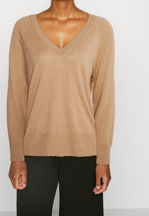 Frau trägt einen beigen Langarm-V-Ausschnitt-Pullover kombiniert mit schwarzen Hosen vor einem einfarbigen hellen Hintergrund.