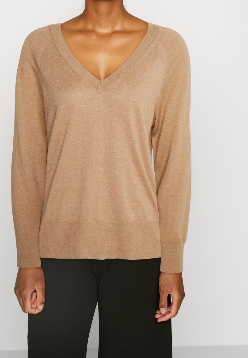 Femme portant un pull beige à manches longues avec col en V, associé à un pantalon noir sur un fond clair uni.