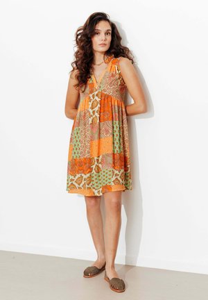 Femme aux longs cheveux bouclés portant une robe sans manches à motifs patchwork orange et vert et des chaussures marron sans lacets, debout contre un mur blanc.