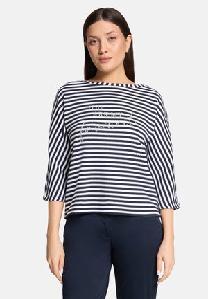 Frau trägt ein marineblau-weiß gestreiftes Langarmshirt mit Text und eine dunkelblaue Hose, steht vor einfarbigem Hintergrund.