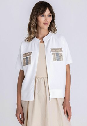 Femme portant une veste blanche à manches courtes avec fermeture éclair et poches avant décorées par-dessus une robe beige, debout devant un fond uni.