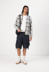 Camisa de cuadros en verde y azul, camiseta blanca de cuello redondo, pantalones cortos de cargo oscuros con bolsillos laterales y zapatillas beige con suelas de goma.