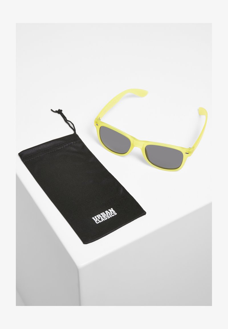 Dzelteni saulesbrilles ar pelēkiem lēcēm, taisnstūra forma. Ietver melnu maisiņu ar "URBAN CLASSICS" logotipu baltā krāsā.