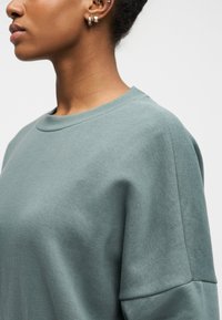 Προφίλ ενός ατόμου που φοράει μια απαλό πράσινο φούτερ, με σκουλαρίκια σε τρυπημένα αυτιά και ένα μικρό τατουάζ στο λαιμό, σε απλό φόντο.