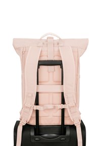 Rosa tygryggsäck med vadderade remmar, rulltoppsdesign och nätdetaljer, fäst på ett svart hjulvagnshandtag.