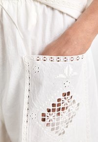 Λευκό ένδυμα από ύφασμα με λεπτομερείς δαντελένιες λεπτομέρειες και κεντητές κοψίματα στην τσέπη, επιδεικνύοντας γεωμετρικά σχέδια και λουλουδένιες προσθήκες.