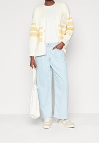 Cardigan a maglia chiara con righe gialle e toppe, abbinato a jeans azzurri boyfriend e sneakers beige, mentre tiene una borsa a tracolla bianca.