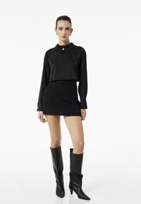 Schwarze Bluse mit langen Ärmeln, Kragen und Knopfdetail, kombiniert mit einem taillierten schwarzen Minirock und knielangen schwarzen Stiefeln mit Absatz.