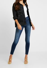 Chaqueta de mezclilla negra con costuras blancas contrastantes, combinada con jeans ajustados azules con dobladillos deshilachados y sandalias amarillas con tiras.