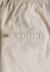 Beige kangas, jossa on sileä pinta, takatasku ja ommeltu yksityiskohtia. Sisältää metallisen logo-merkin taskussa.