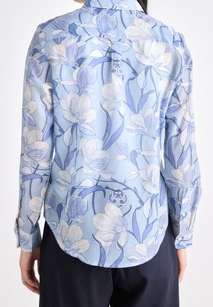 Chemise bleu clair à manches longues avec motif floral blanc et bleu portée par une personne de dos, associée à un pantalon foncé.