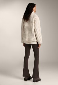 Strukturierter, cremefarbener Pullover mit Fischgrätmuster, oversized Schnitt, hohem Kragen und geripptem Saum, kombiniert mit dunklen Schlaghosen und Stiefeln.