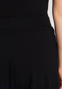 Combinaison noire en tissu lisse et extensible, taille ajustée, avec un détail en dentelle festonnée le long de l'ourlet. Design simple pour un port polyvalent.