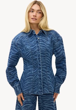 Femme portant une veste en jean bleue et un pantalon à rayures zébrées foncées, debout les mains le long du corps sur fond blanc.