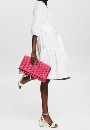 Woven pink clutch bag with a zip closure, held in a hand. Model wearing a white dress and strappy white heels, showing elegant shapes and textures.