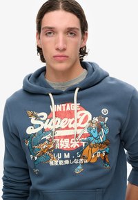 Sudadera azul con un gráfico frontal que presenta dragones y guerreros. Tiene cierre con cordón y una textura suave. Logotipo de la marca presente.