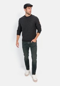 Dunkelgrauer strukturierter Pullover, getragen mit dunklen Jeans. Hellgraue Sneakers mit beigen Sohlen. Schwarze Schirmmütze auf dem Kopf. Schlichtes, modernes Design.