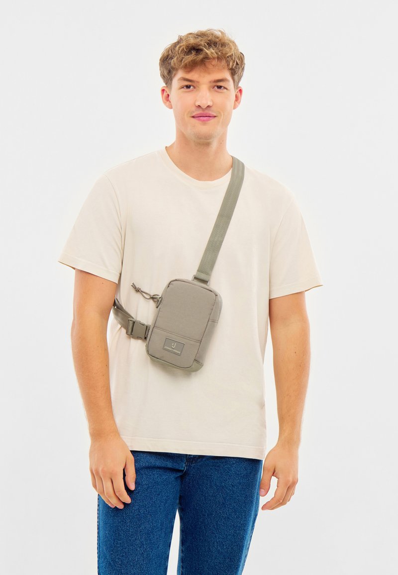 Ung mand iført en beige t-shirt og blå jeans med en grå crossbody taske spændt over brystet mod en ensfarvet baggrund.