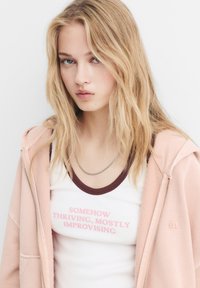 Roze zip-up hoodie over een witte tanktop met bruine accenten en roze tekst die leest "SOMEHOW THRIVING, MOSTLY IMPROVISING". Zilveren ketting.