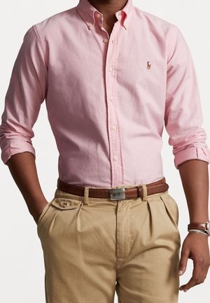 Homme portant une chemise rose clair à boutons avec manches retroussées, pantalon beige plissé, ceinture en cuir marron et montre-bracelet avec bracelets.