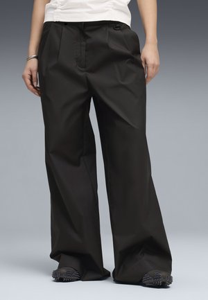 Person wearing wide-leg black trousers and black shoes with textured soles, standing against a plain gray background.