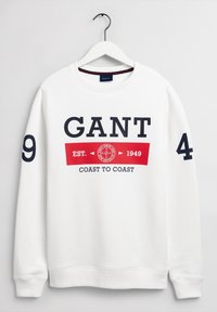 Sudadera blanca con texto en azul marino y detalles en rojo. Incluye gráficos de "GANT", "EST. 1949" y "COAST TO COAST". Puños y cuello acanalados.