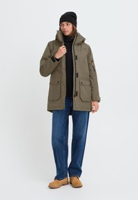 Olivenfarget parka med hette, med flere frontlommer, svart maskinvare, svart genser under, blå jeans og brune støvler.