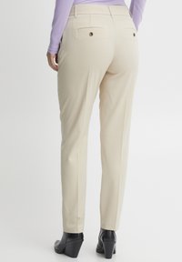 Pantaloni beige su misura con una vestibilità dritta, dotati di due tasche posteriori e dettagli con bottoni marroni. Indossati con un top viola e scarpe col tacco nere.